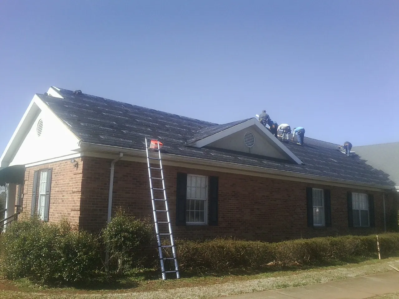 Crew tearing shingles off a brick ranch roof with a ladder set — Emergency Roof Repair work by our crew in Hoschton