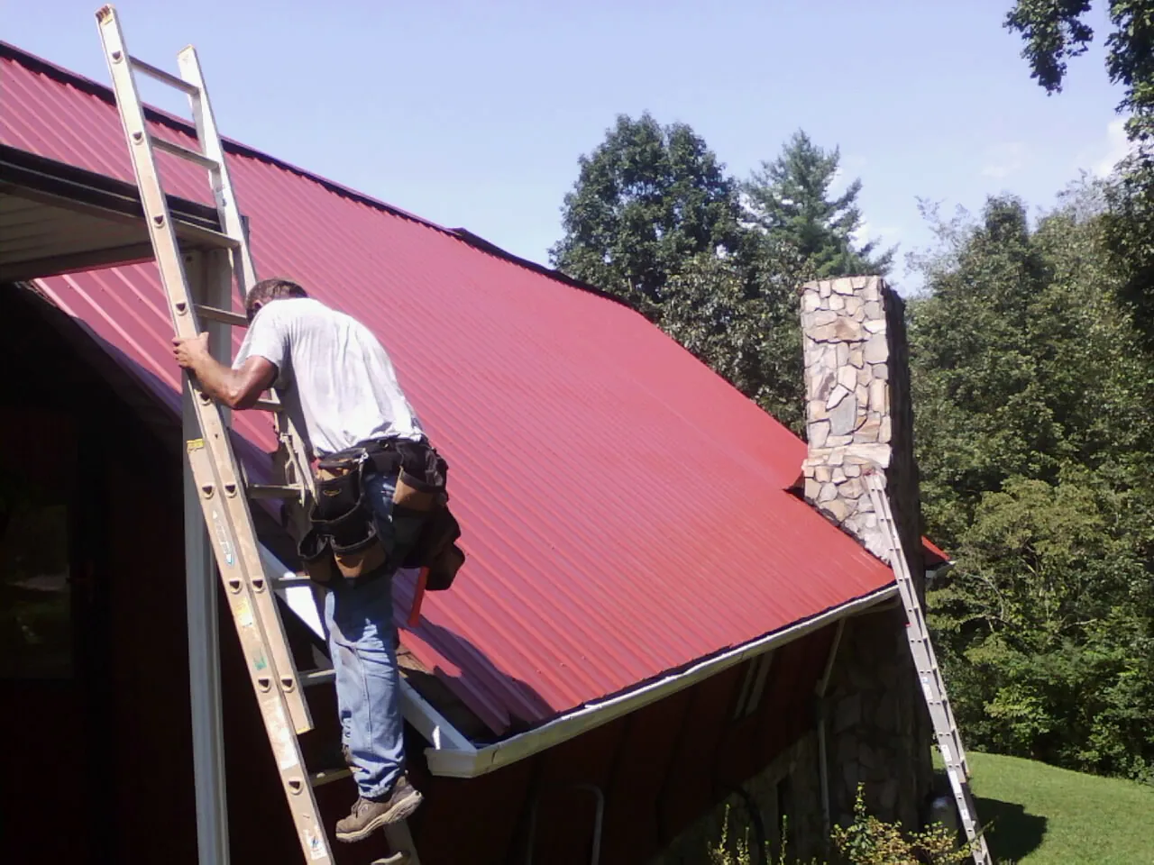 Roofer on a ladder beside a red metal roof and stone chimney — Soffit & Fascia Repair by Roofing Brothers in Hoschton